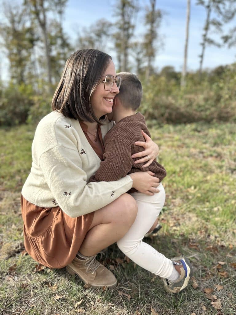 Portrait de profil de Charline Pancelot, naturopathe Angers en robe marron, accroupi et faisant un sourire dans la forêt et tenant un enfant contre son corps.