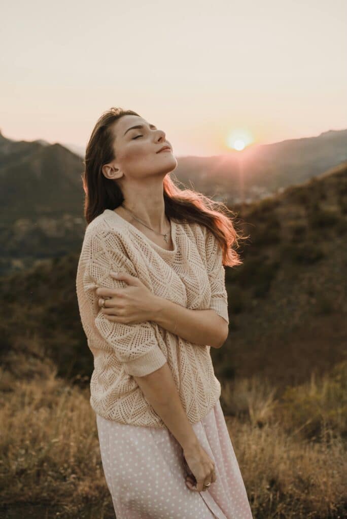 Femme dans la nature devant un soleil couchant se tenant un bras et regardant le ciel avec soulagement.